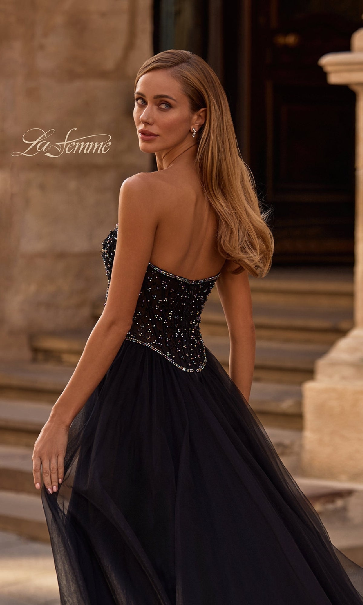 A woman in the La Femme 32856 strapless long black prom ball gown with a beaded bodice stands outdoors, turning to look over her shoulder.